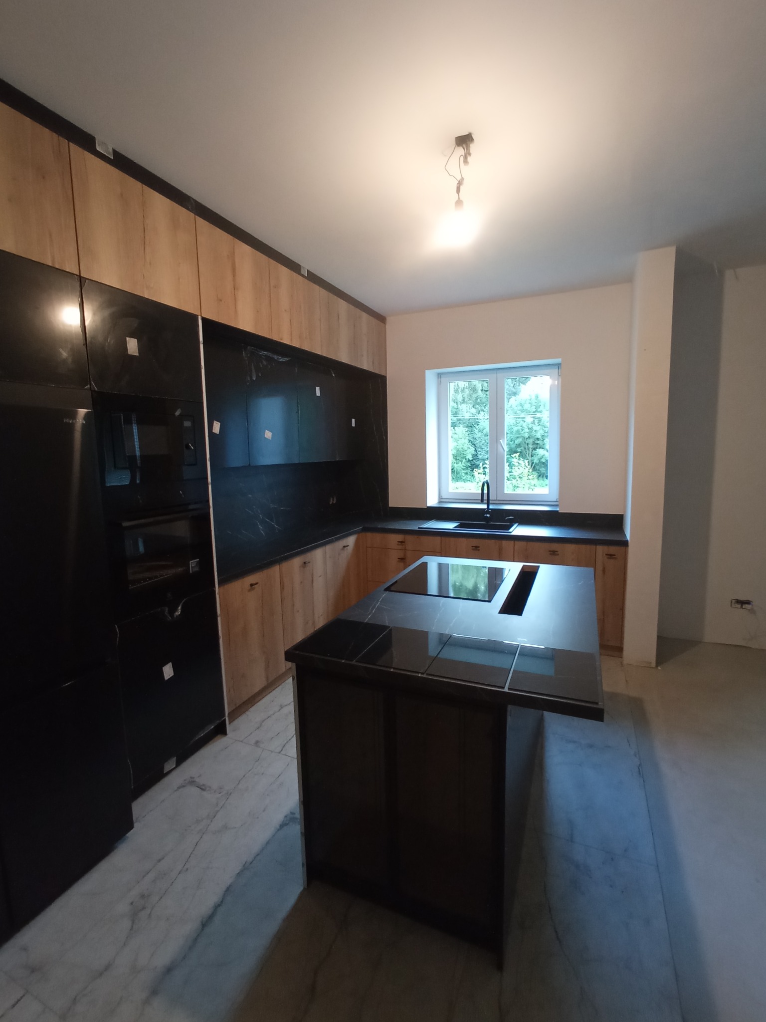 Nowoczesna kuchnia z wyspą: drewniane szafki, czarne blaty i marmurowa podłoga. Widok na okno z zielenią. Minimalistyczne oświetlenie sufitowe.