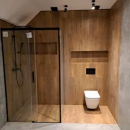 Nowoczesna łazienka z kabiną prysznicową typu walk-in, drewnopodobnymi płytkami na ścianach i podłodze, oraz podwieszaną miską WC. Oświetlenie punktowe.