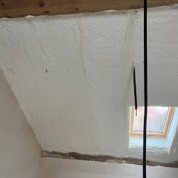 Poddasze po ociepleniu pianką PUR, widoczne drewniane belki stropowe i okno dachowe. Biała, nierówna powierzchnia pianki pokrywa skośne ściany i sufit.