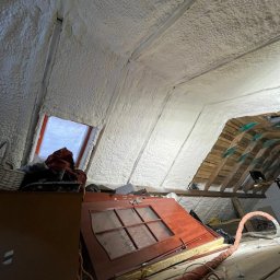 Poddasze po ociepleniu pianką PUR, widoczne białe wypełnienie między krokwiami, okno dachowe i fragment drewnianej konstrukcji dachu. Częściowo widoczne przedmioty na podłodze.