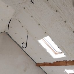 Poddasze po ociepleniu pianką PUR, widoczne okno dachowe i przewody elektryczne. Biała, porowata struktura pianki pokrywa skośne powierzchnie.