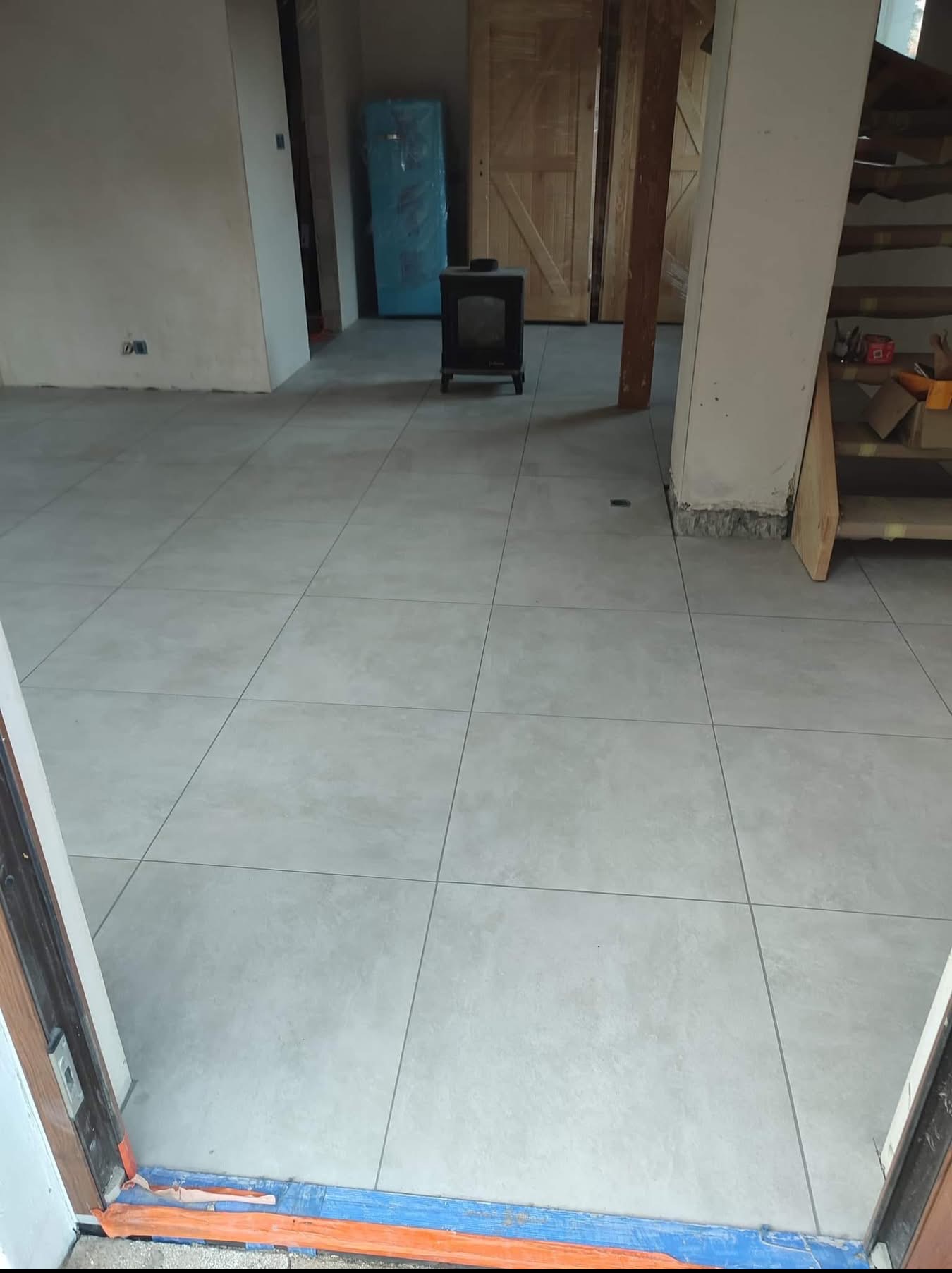 Jasne, kwadratowe płytki podłogowe ułożone w nowym wnętrzu. W tle piec wolnostojący, drzwi z surowego drewna i schody. Widok z progu.