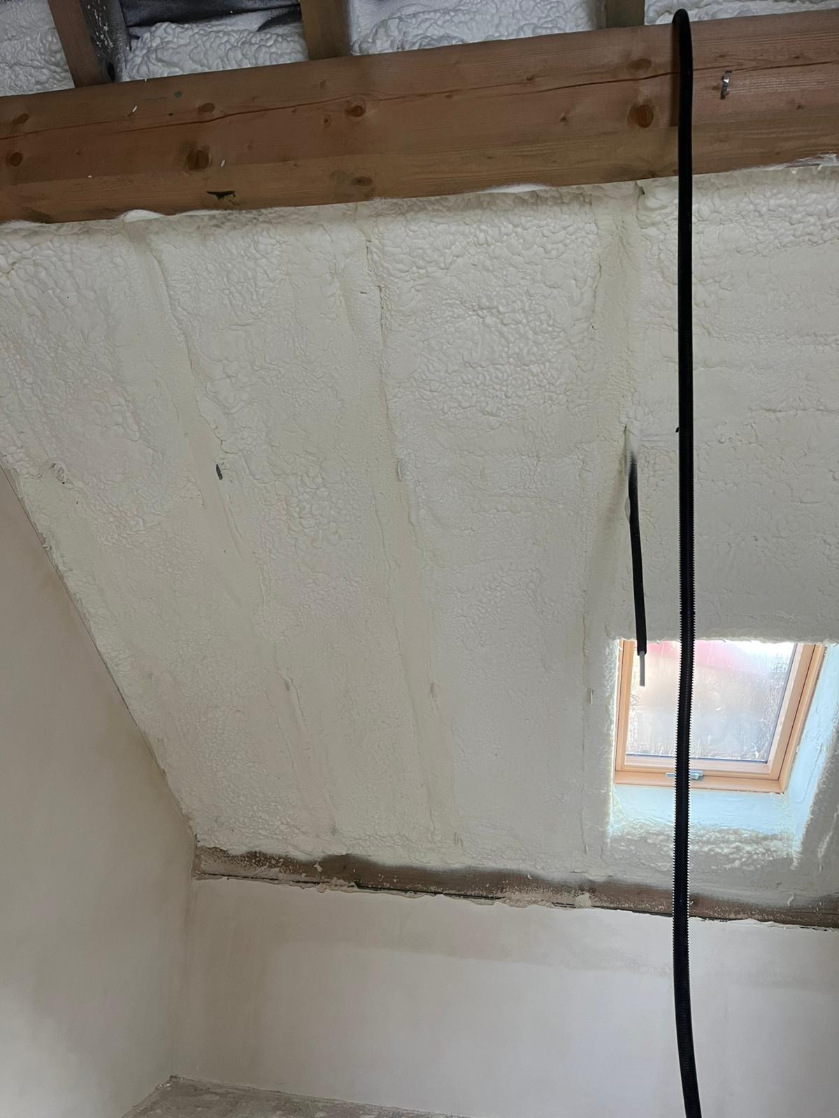 Poddasze po ociepleniu pianką PUR, widoczne drewniane belki stropowe i okno dachowe. Biała, nierówna powierzchnia pianki pokrywa skośne ściany i sufit.