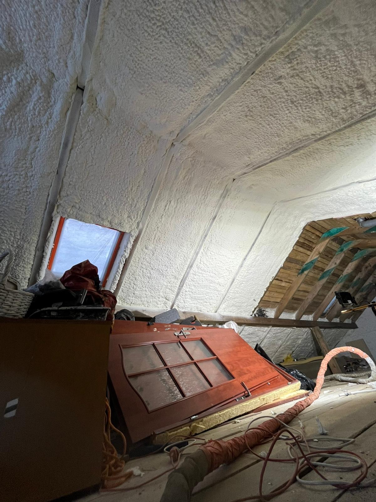 Poddasze po ociepleniu pianką PUR, widoczne białe wypełnienie między krokwiami, okno dachowe i fragment drewnianej konstrukcji dachu. Częściowo widoczne przedmioty na podłodze.