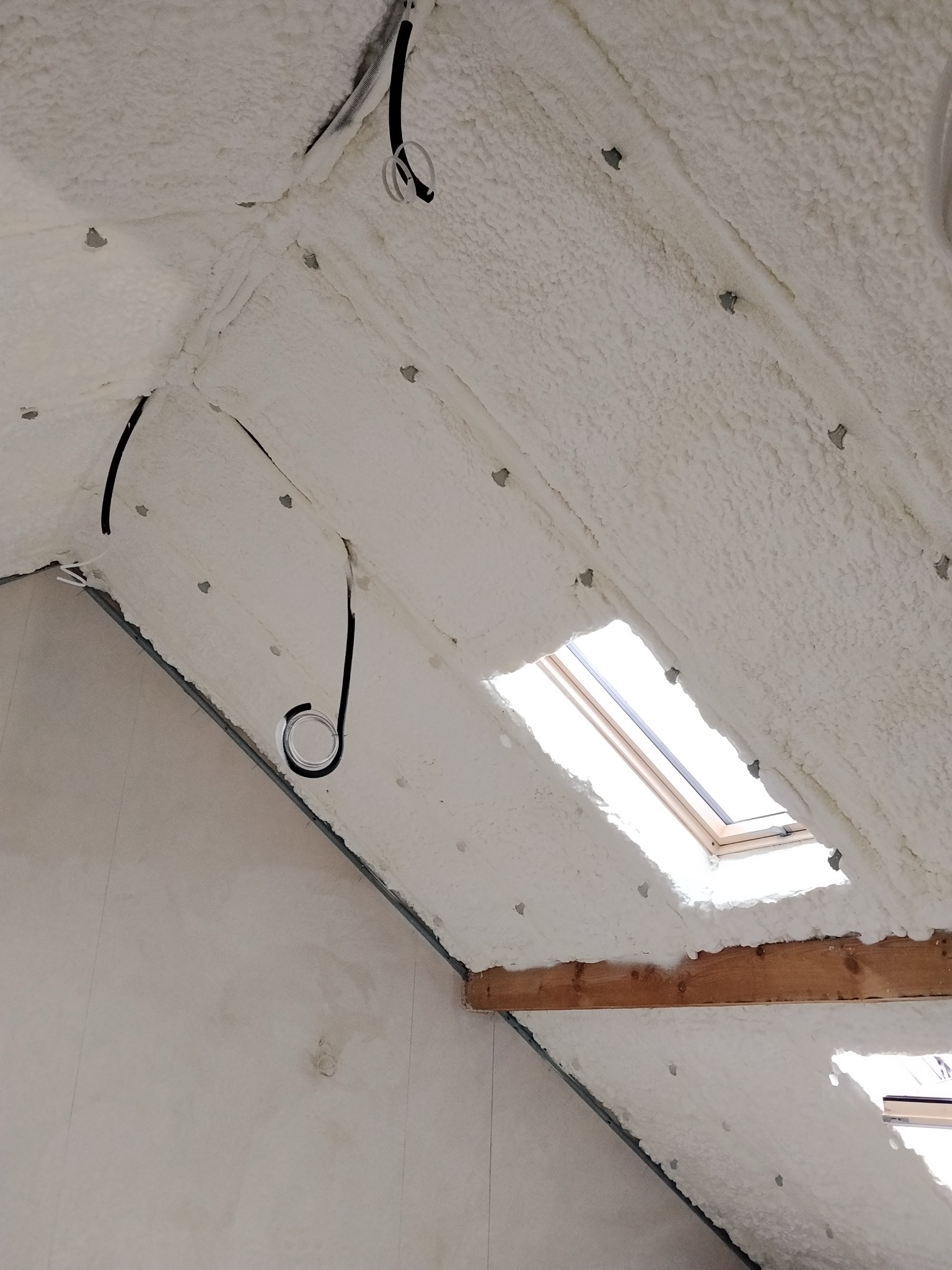 Poddasze po ociepleniu pianką PUR, widoczne okno dachowe i przewody elektryczne. Biała, porowata struktura pianki pokrywa skośne powierzchnie.