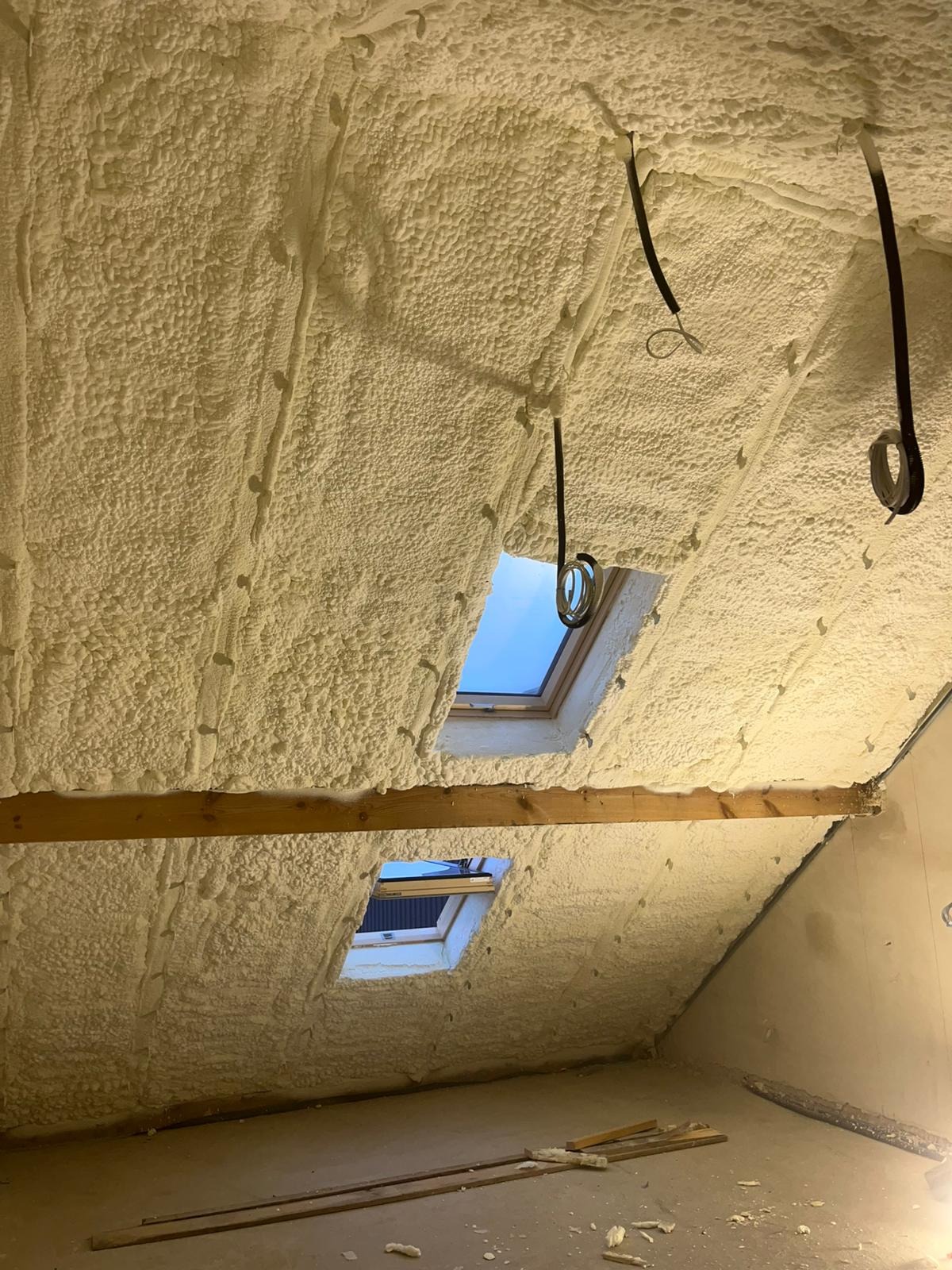 Poddasze po ociepleniu pianką PUR, widoczne okna dachowe i drewniane belki. Kabel elektryczny zwisa z piany. Na podłodze resztki piany i drewniane listwy.