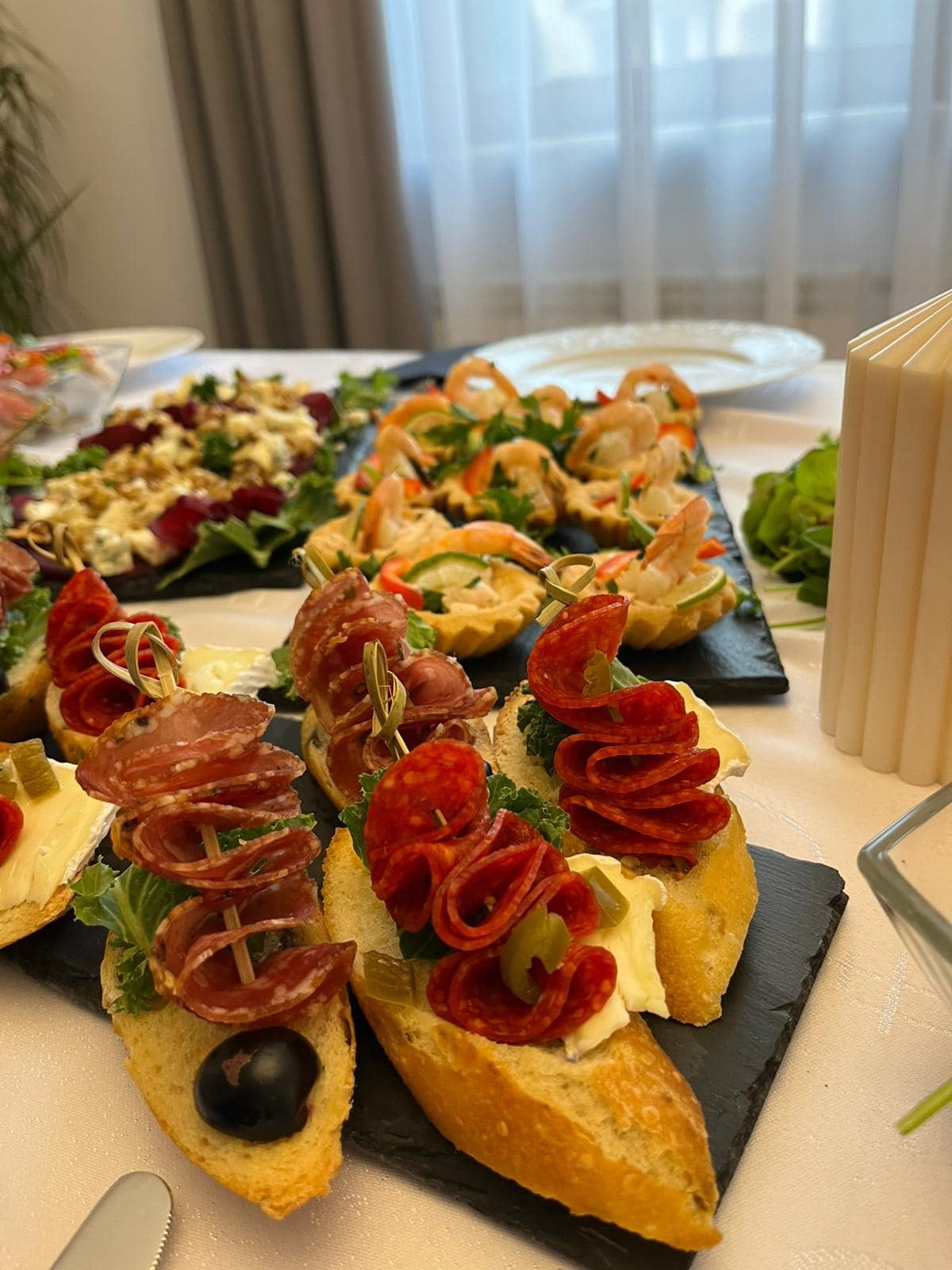 Elegancko podane przekąski cateringowe: kanapeczki z salami, tartaletki z krewetkami i sałatka na ciemnych deskach, na białym obrusie, w tle okno z firanką.