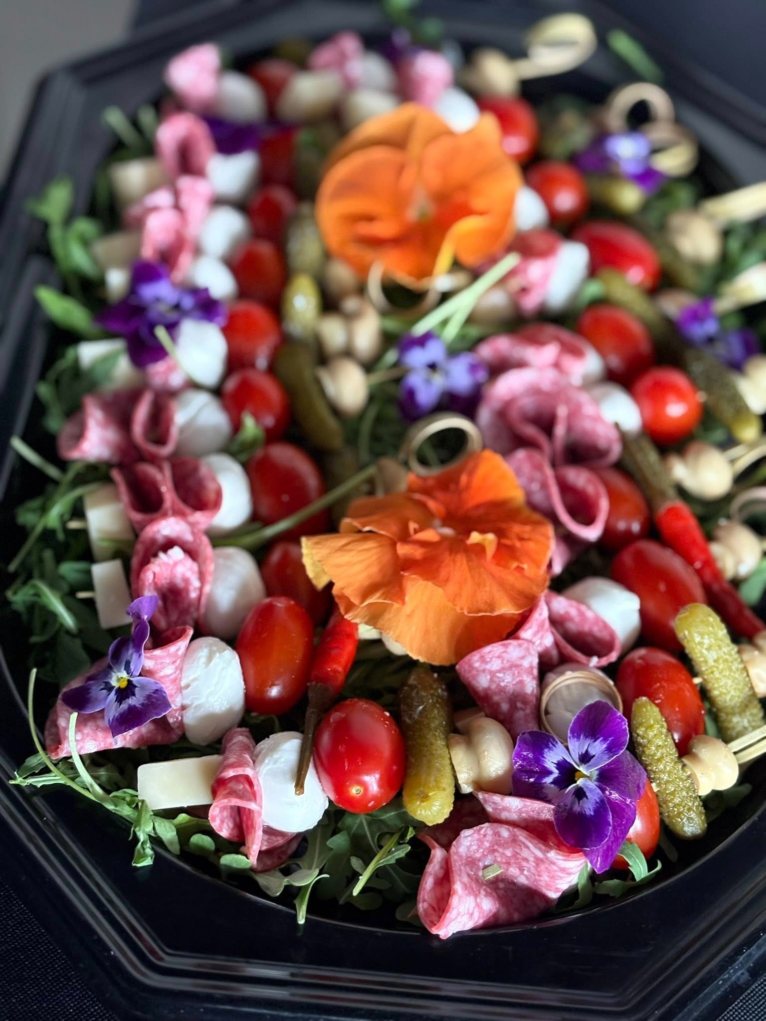 Elegancka taca cateringowa z przekąskami: salami, mini mozzarella, pomidorki, ogórki konserwowe, grzyby i jadalne kwiaty. Idealne na imprezę firmową.