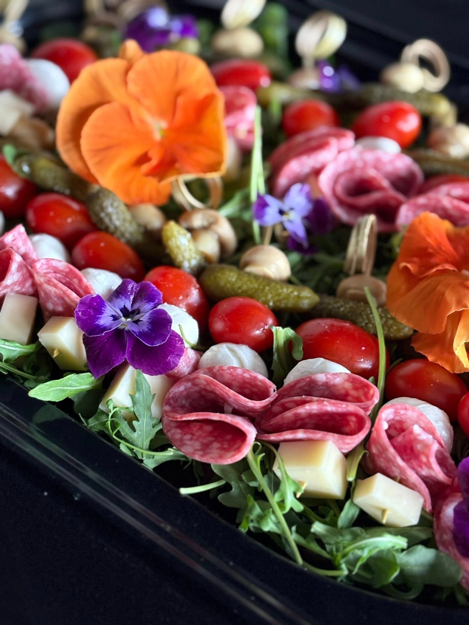 Dekoracyjna taca z przekąskami: salami, ser, pomidorki, ogórki, mozzarella, rukola, ozdobiona jadalnymi kwiatami w odcieniach fioletu i pomarańczy.