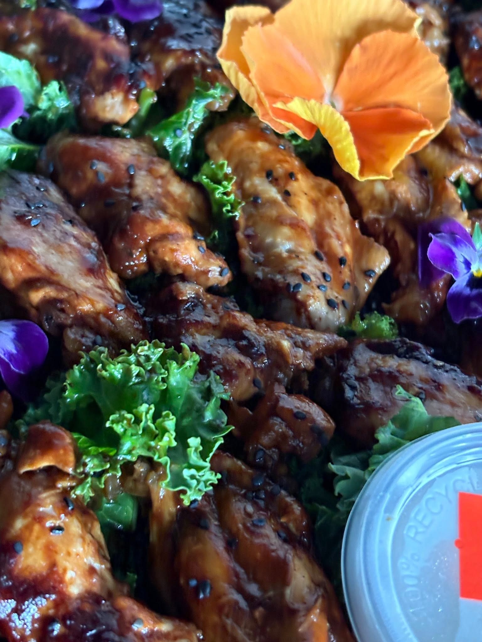 Apetyczne kąski kurczaka w glazurze z sezamem, udekorowane świeżymi kwiatami i zielonymi liśćmi sałaty, gotowe do serwowania w pojemniku.