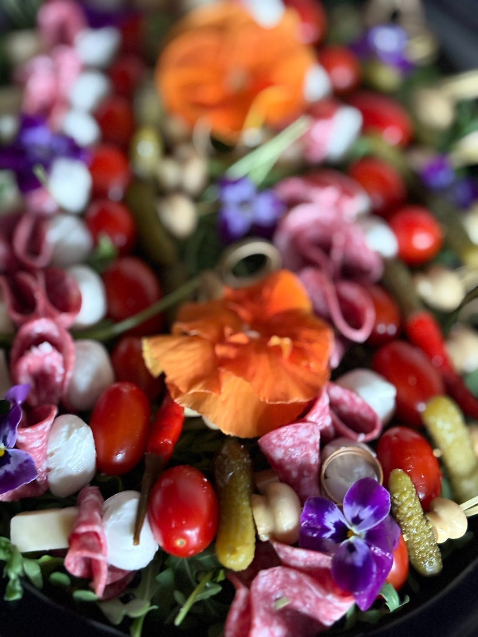 Dekoracyjna kompozycja przekąsek: salami, mini mozzarella, pomidorki koktajlowe, korniszony, grzybki i fiołki na ciemnym tle.