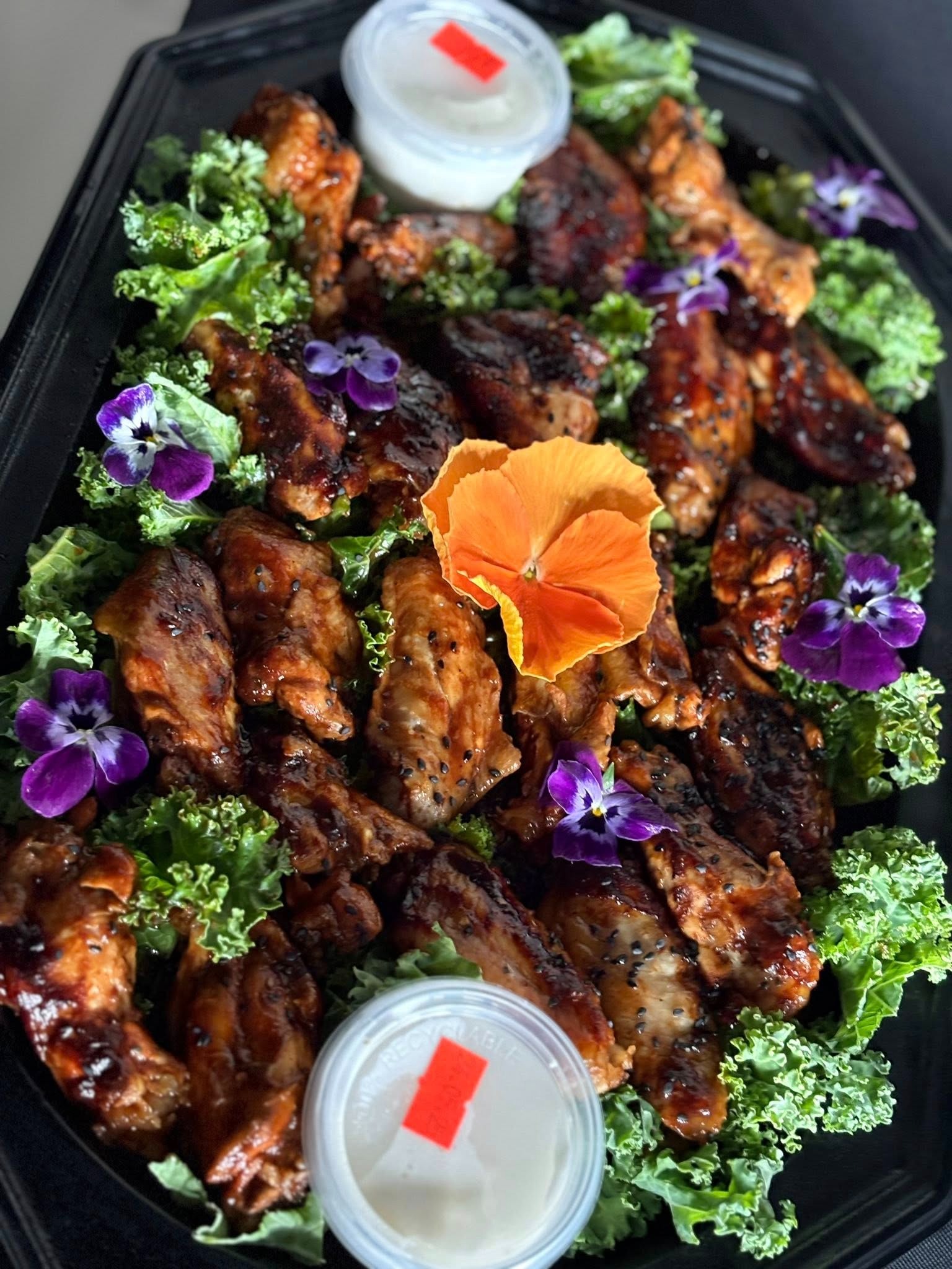 Elegancki catering: błyszczące skrzydełka kurczaka z sezamem, dekorowane jadalnymi kwiatami i sałatą na czarnej tacy, z dwoma pojemnikami z sosem.