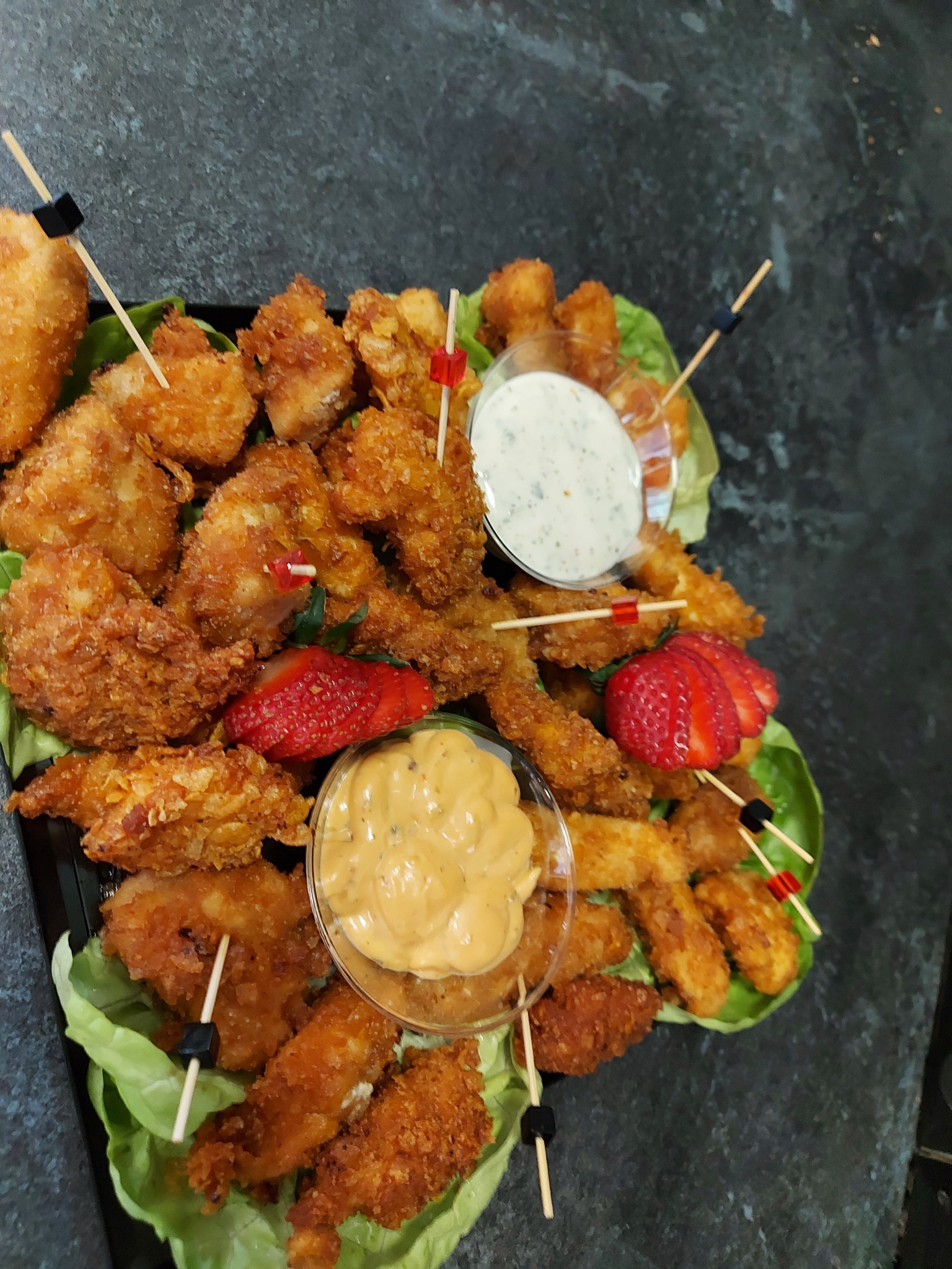 Elegancka taca z przekąskami: chrupiące kąski z kurczaka na sałacie, kolorowe wykałaczki, dwa sosy w szklanych miseczkach i plasterki truskawek. Catering.