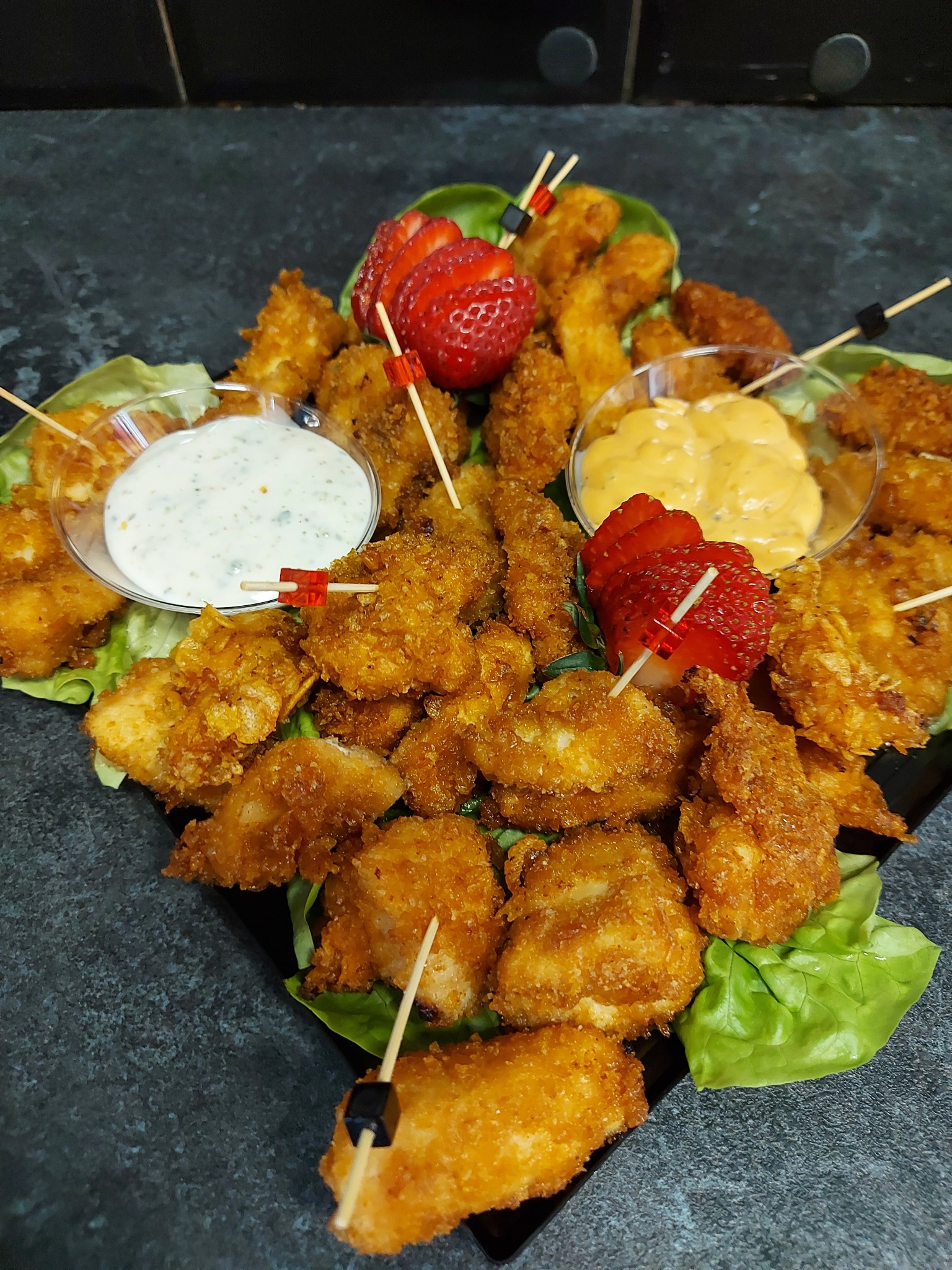 Apetyczne nuggetsy z kurczaka, serwowane z dwoma sosami w szklanych miseczkach i dekorowane świeżymi truskawkami, na czarnym talerzu z zieloną sałatą.