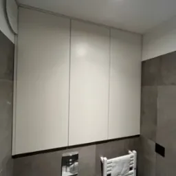 Białe szafki łazienkowe wiszące nad grzejnikiem drabinkowym z ręcznikiem, na tle szarych płytek. Widoczny panel sterujący spłuczką.