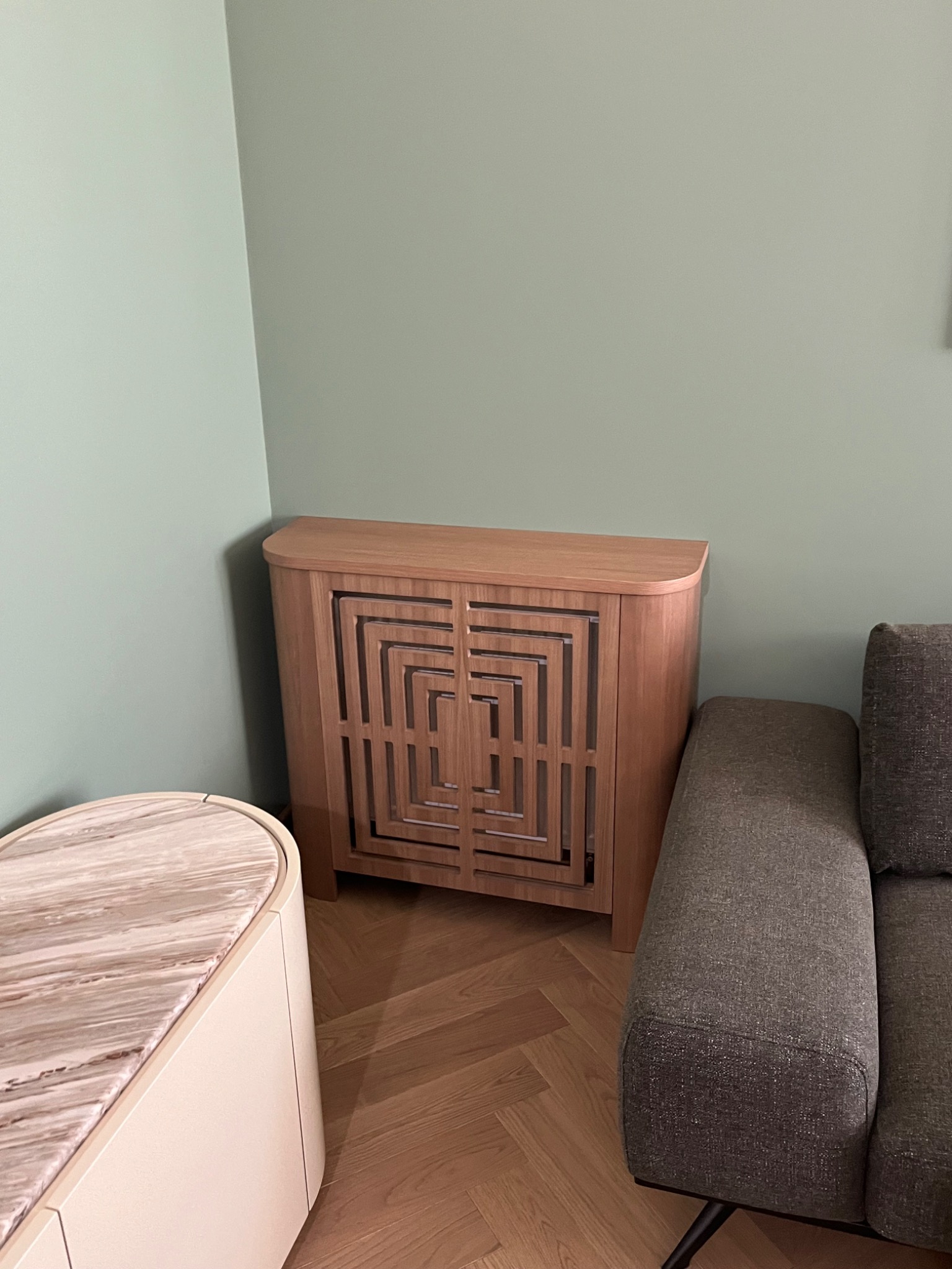 Drewniana szafka z geometrycznym wzorem, stojąca obok sofy i stolika na parkiecie, na tle zielonej ściany. Widok z bliska, oświetlenie naturalne.