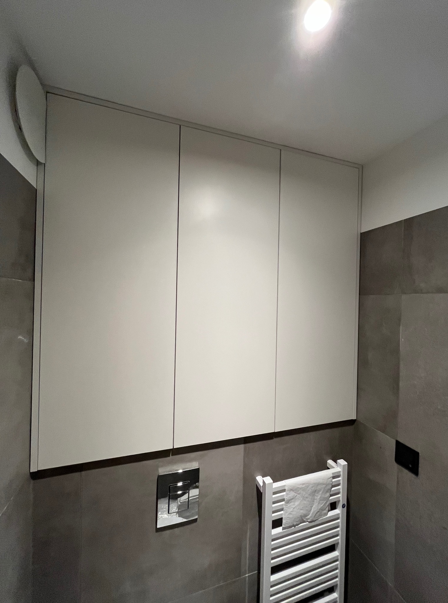 Białe szafki łazienkowe wiszące nad grzejnikiem drabinkowym z ręcznikiem, na tle szarych płytek. Widoczny panel sterujący spłuczką.