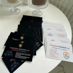 Stos czarnych i białych wizytówek Telekom Ekspert AS na białym stoliku obok świec. Eleganckie, minimalistyczne wzornictwo. Detal: logo z błyskawicą.