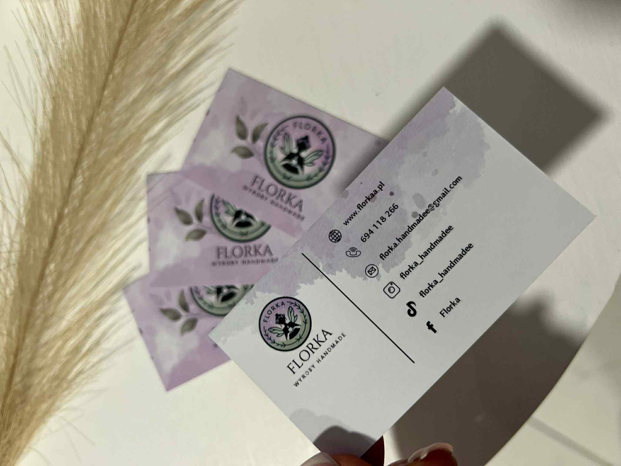 Eleganckie wizytówki firmy Florka Wyroby Handmade z logo i danymi kontaktowymi, ułożone na białym tle z dekoracyjnym źdźbłem trawy pampasowej.