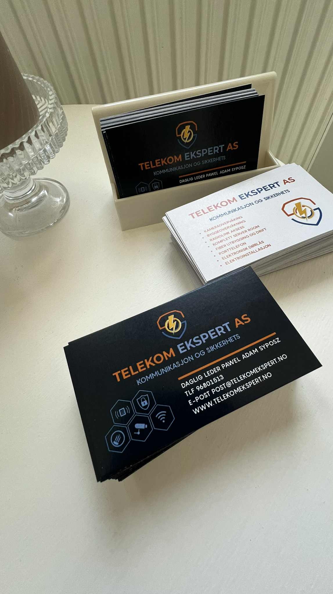 Stos wizytówek firmy Telekom Ekspert AS, czarne i białe, leżące na białym stole obok szklanego wazonu. Wizytówki prezentują logo i dane kontaktowe firmy.