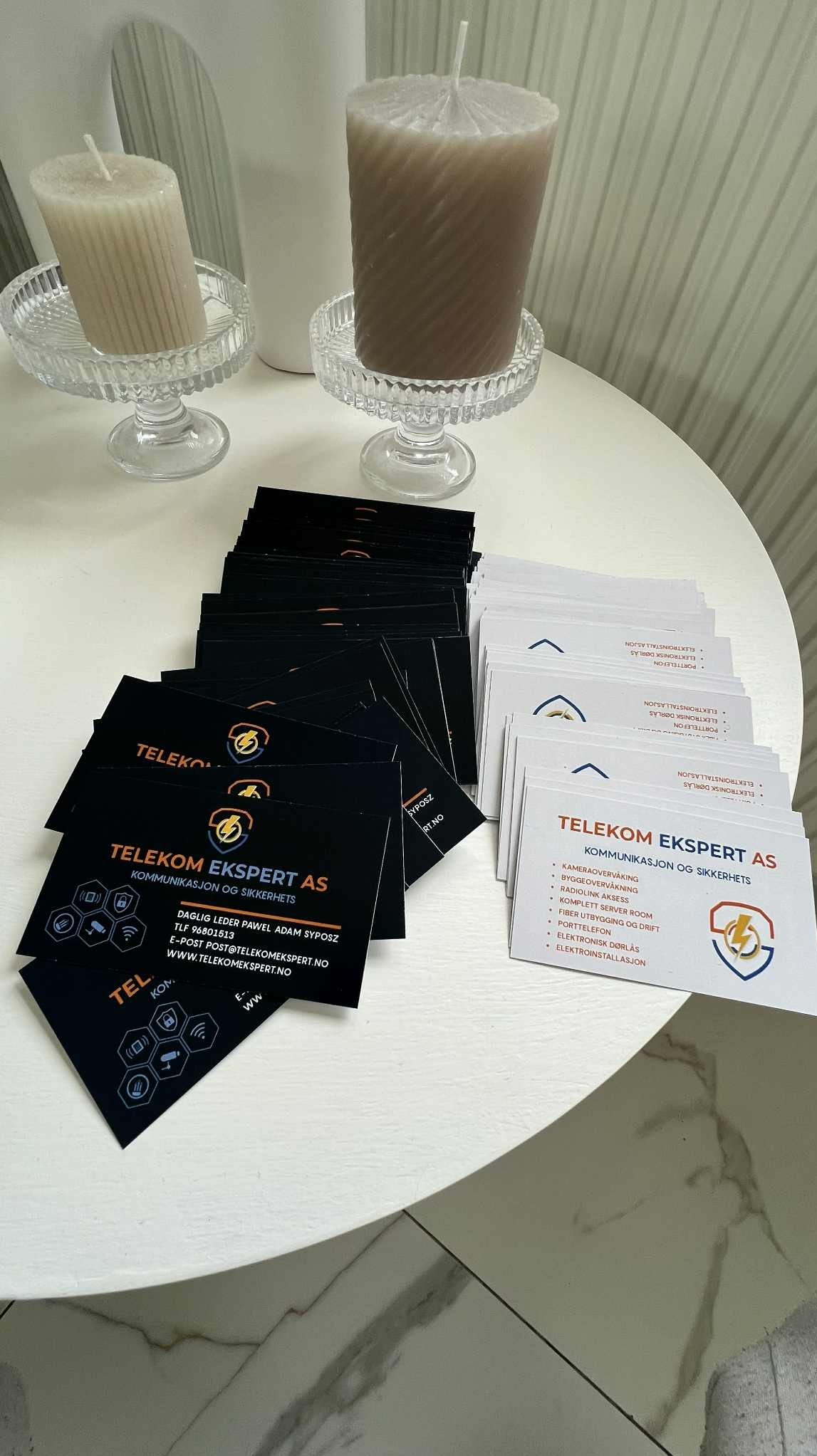 Stos czarnych i białych wizytówek Telekom Ekspert AS na białym stoliku obok świec. Eleganckie, minimalistyczne wzornictwo. Detal: logo z błyskawicą.