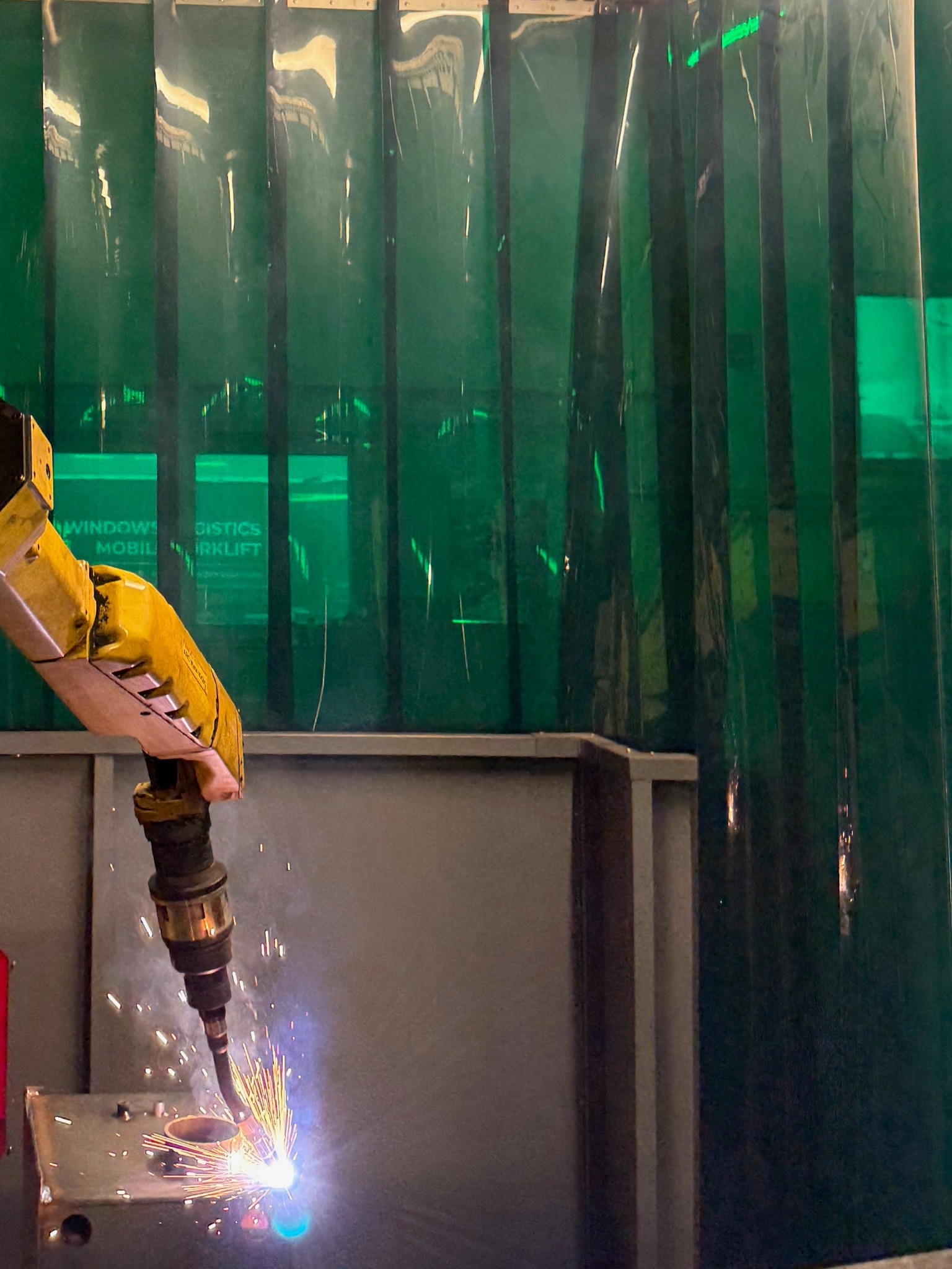 Żółty robot spawalniczy w akcji, iskry i jasne światło podczas spawania metalowego elementu, zielona kurtyna spawalnicza w tle.