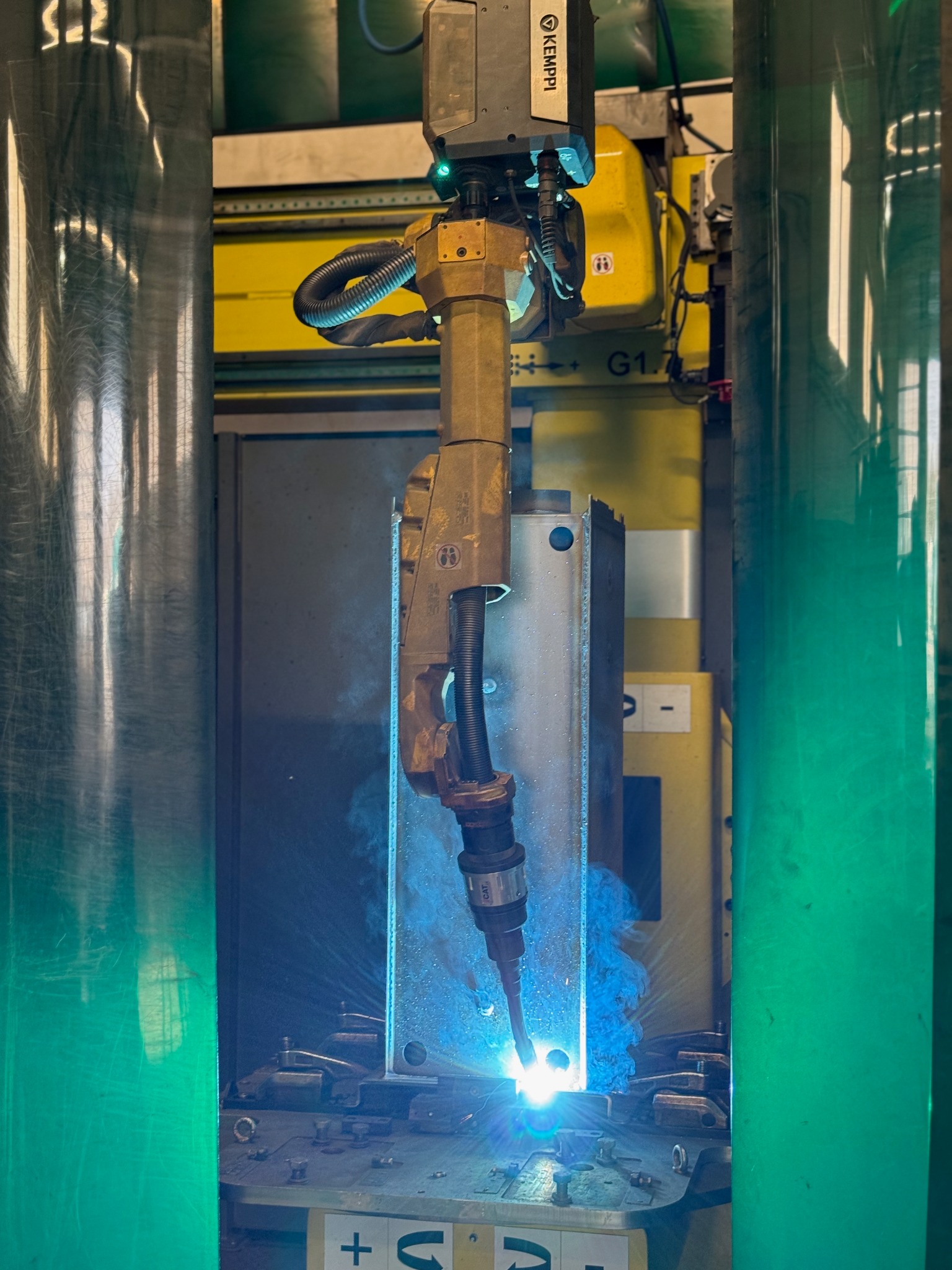 Zautomatyzowane stanowisko spawalnicze z robotem Kemppi w trakcie pracy. Widoczne iskry i dym podczas spawania metalowego elementu w osłonie gazów.
