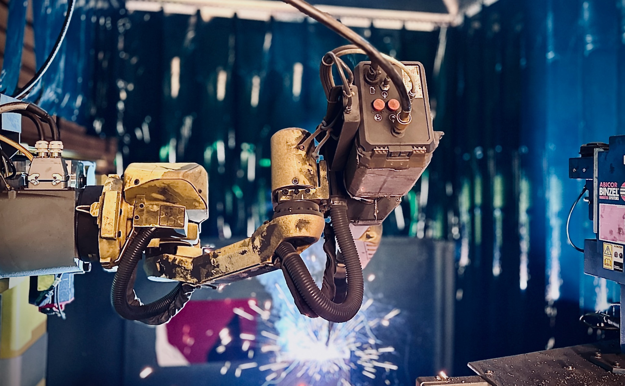 Zautomatyzowane stanowisko spawalnicze z robotem przemysłowym w trakcie pracy, iskry i niebieskie kurtyny ochronne w tle. Widoczne elementy sterujące i przewody.