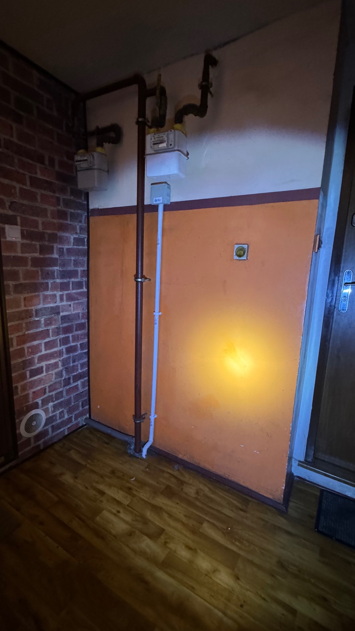 Instalacja gazowa w rogu pomieszczenia z ceglaną ścianą i pomarańczową ścianą z rurami oraz licznikiem. Widoczne korytka i puszki instalacyjne.