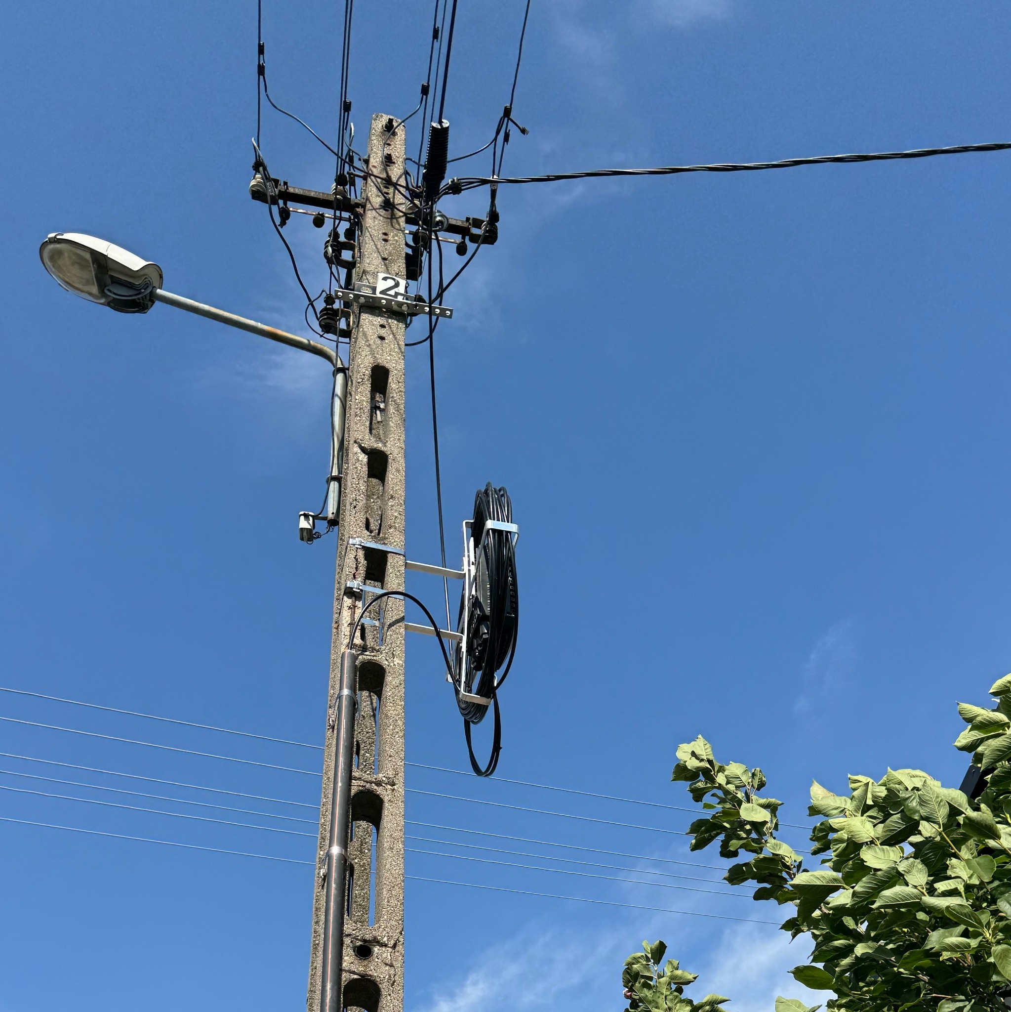Betonowy słup telekomunikacyjny z zamontowanym kablem światłowodowym na stelażu, widok z dołu na tle błękitnego nieba. Widoczna latarnia uliczna i zieleń.