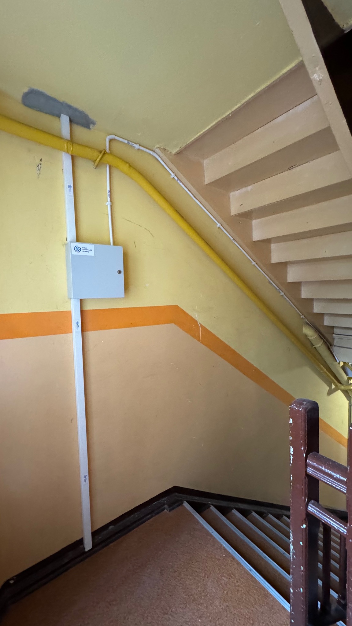 Instalacja rur, korytka i puszki w klatce schodowej. Żółte i białe rury prowadzone wzdłuż ściany, szara skrzynka na ścianie, widoczne schody i fragment poręczy.