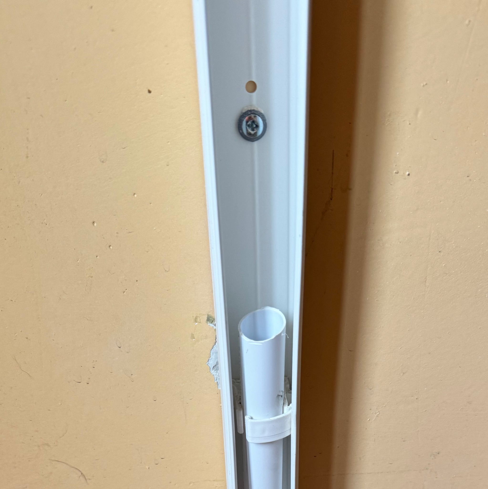Zamocowana biała rura w korytku przy ścianie. Widoczny fragment ściany w kolorze beżowym oraz śruba mocująca koryto. Instalacja w trakcie montażu.