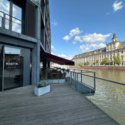 JACEK PIJAR BUDMASTER - Nowoczesny budynek z szarymi żaluzjami zewnętrznymi, widok na rzekę i zabytkowy budynek w tle. Ujęcie z perspektywy tarasu z drewnianą podłogą i szklanymi barierkami.