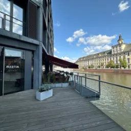 Nowoczesny budynek z szarymi żaluzjami zewnętrznymi, widok na rzekę i zabytkowy budynek w tle. Ujęcie z perspektywy tarasu z drewnianą podłogą i szklanymi barierkami.