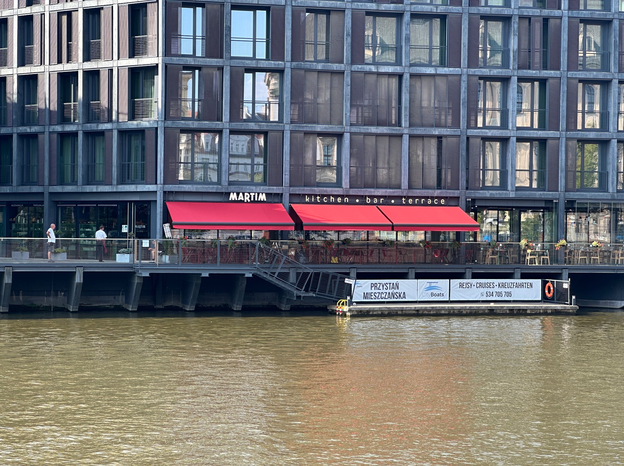 Budynek z żaluzjami fasadowymi w kolorze grafitu, czerwone markizy nad restauracją z widokiem na rzekę. Okna odbijają zabudowę miejską.