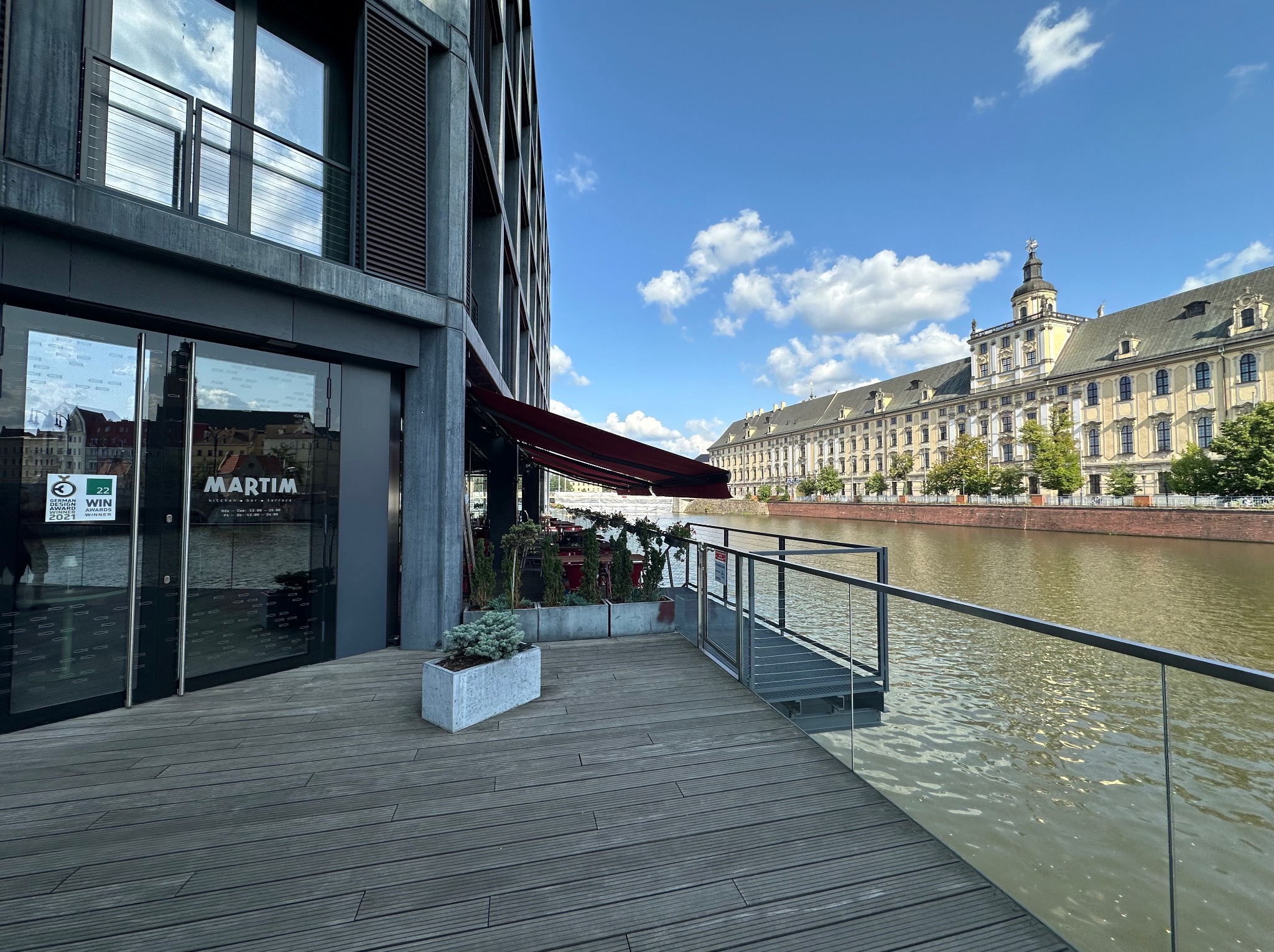 Nowoczesny budynek z szarymi żaluzjami zewnętrznymi, widok na rzekę i zabytkowy budynek w tle. Ujęcie z perspektywy tarasu z drewnianą podłogą i szklanymi barierkami.