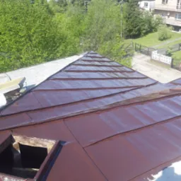 Świeżo pomalowany, brązowy dach z blachy na tle zabudowy mieszkalnej i zieleni, widoczny otwór okienny w dachu.