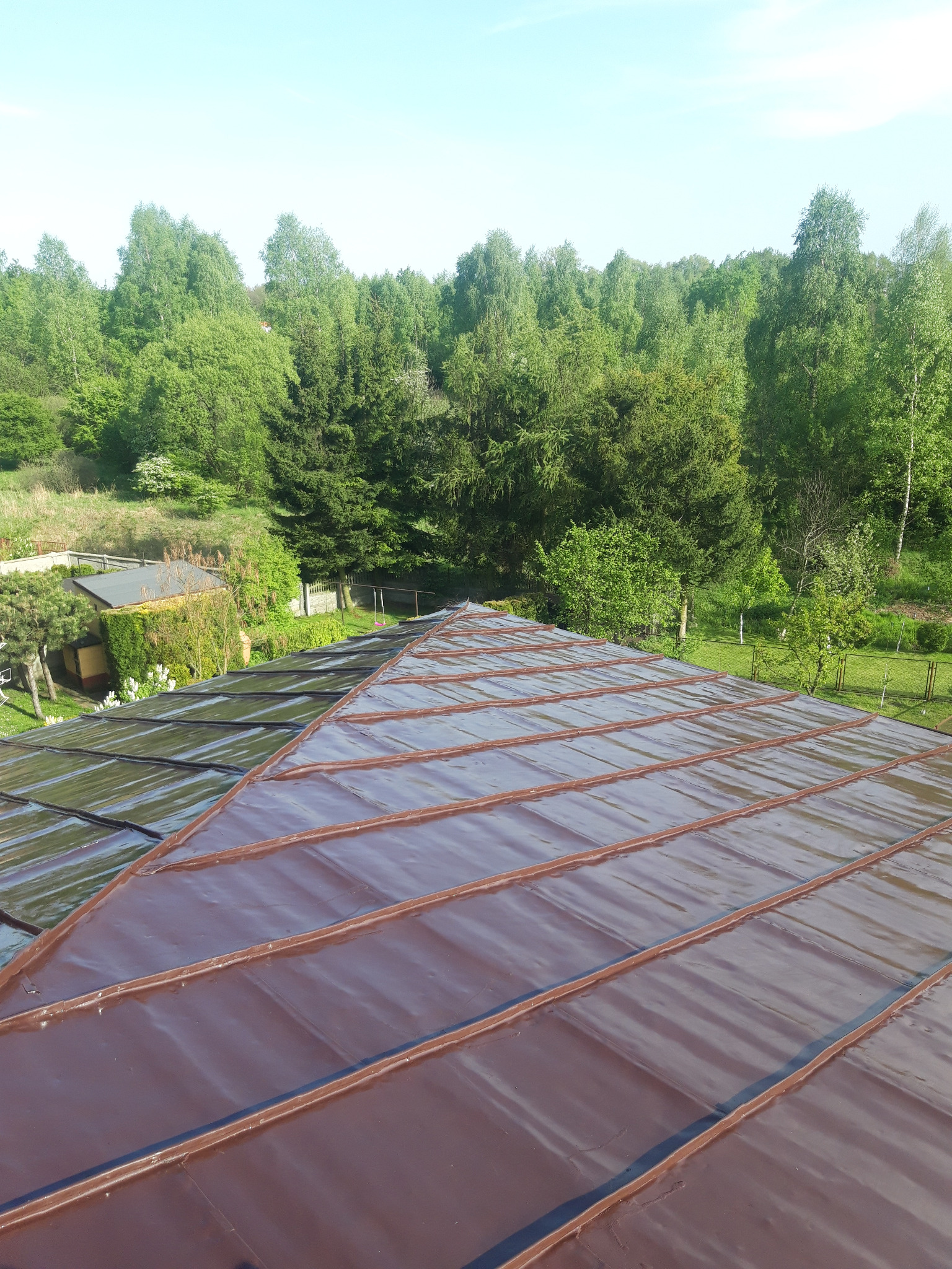 Widok z góry na fragment dachu z brązowego metalu z widoczną obróbką blacharską, w tle zieleń drzew i błękitne niebo.