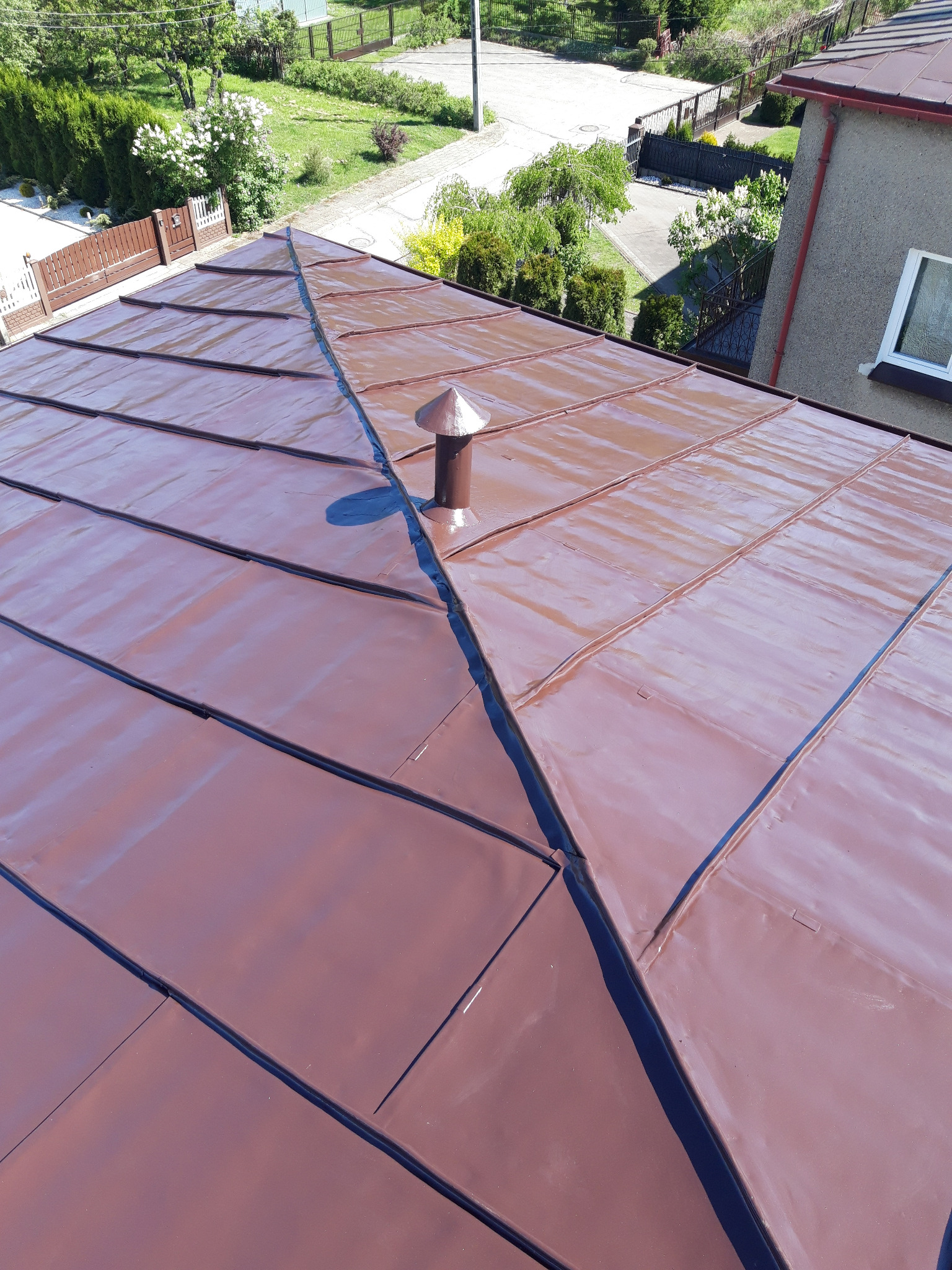 Ciemnobrązowe blaszane pokrycie dachowe z kominkiem wentylacyjnym, widok z góry na tle zieleni ogrodu.