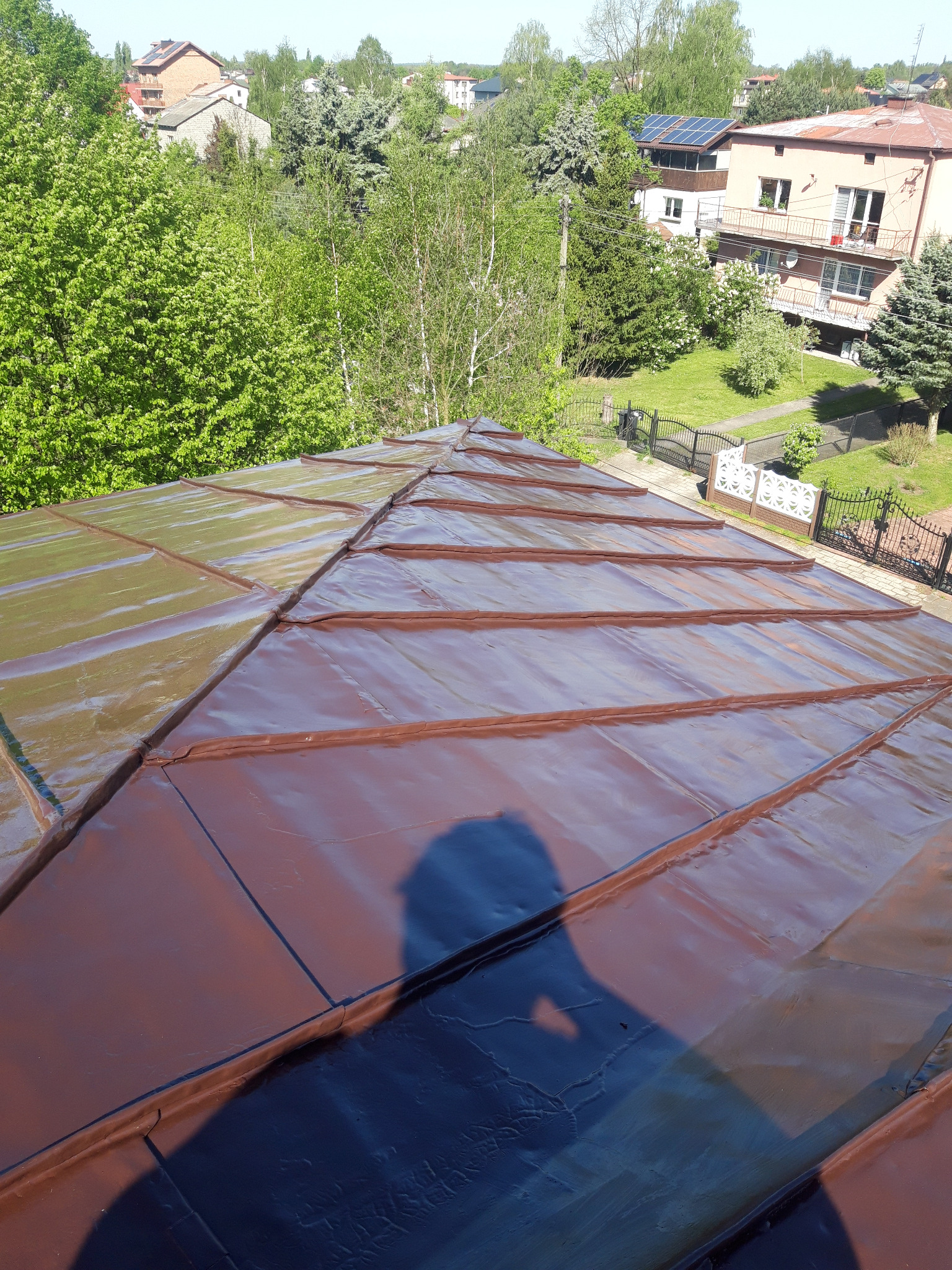 Ciemnobrązowy dach z blachy na tle zieleni drzew i zabudowań, cień osoby fotografującej widoczny na powierzchni dachu.
