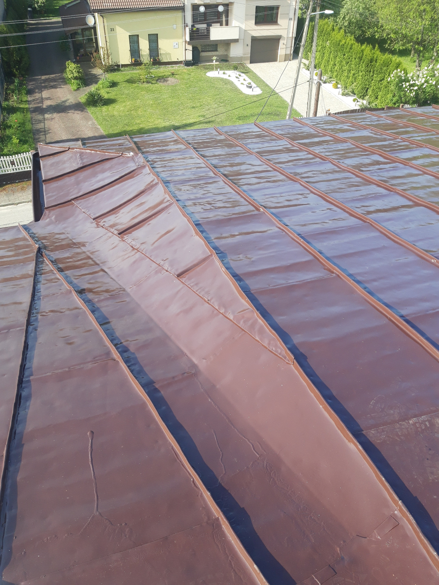 Błyszczące, brązowe blaszane pokrycie dachowe z widocznymi łączeniami i fragmentem dachu sąsiedniego budynku w tle.