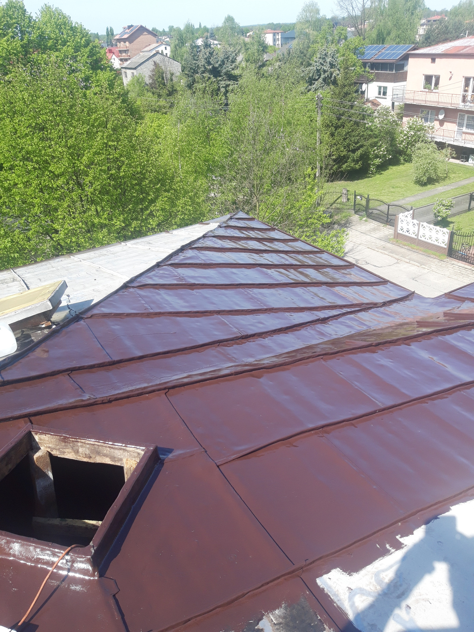 Świeżo pomalowany, brązowy dach z blachy na tle zabudowy mieszkalnej i zieleni, widoczny otwór okienny w dachu.