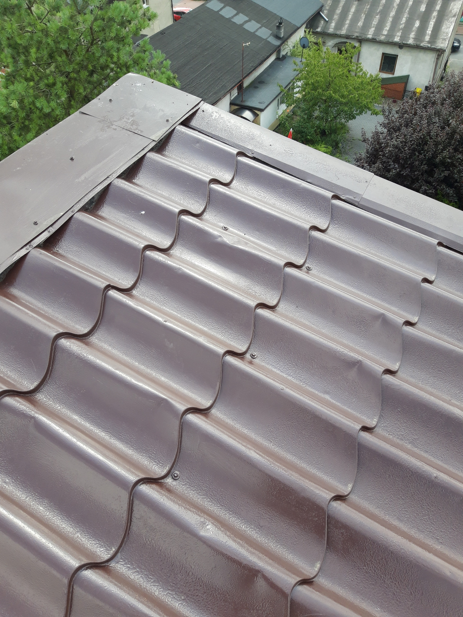 Brązowe, metalowe pokrycie dachowe z widocznymi śrubami mocującymi, obróbka blacharska krawędzi dachu, widok z góry na budynki i zieleń w tle.