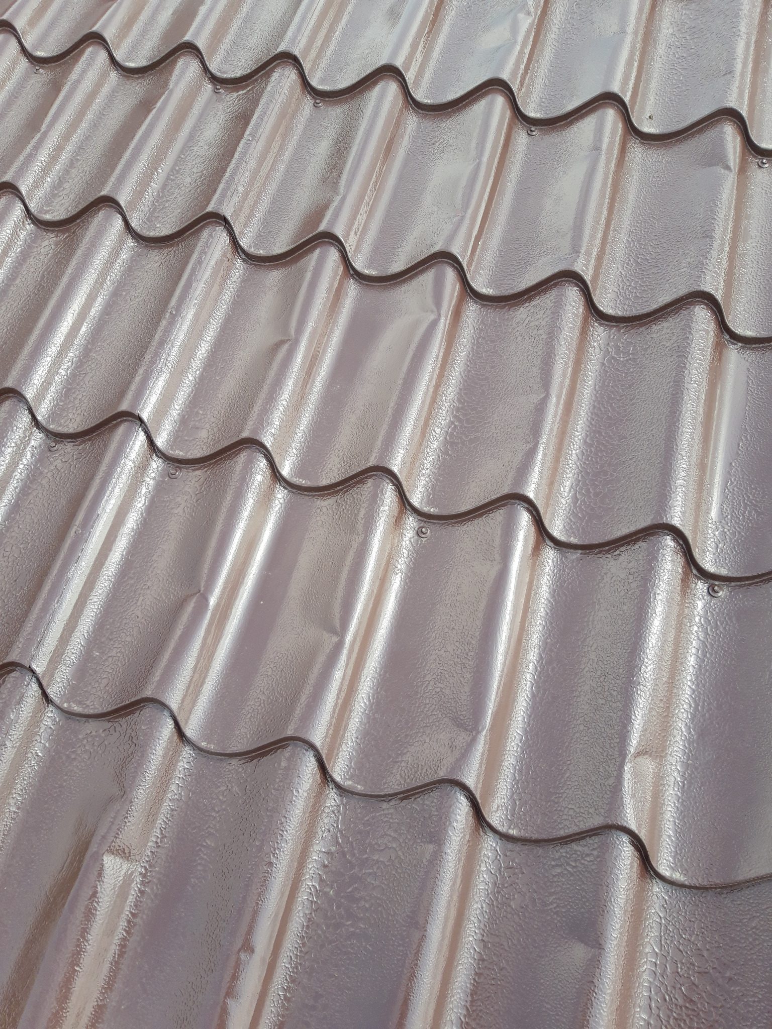 Błyszczące, brązowe panele dachowe z wyraźną teksturą i widocznymi mocowaniami, ułożone w falisty wzór.