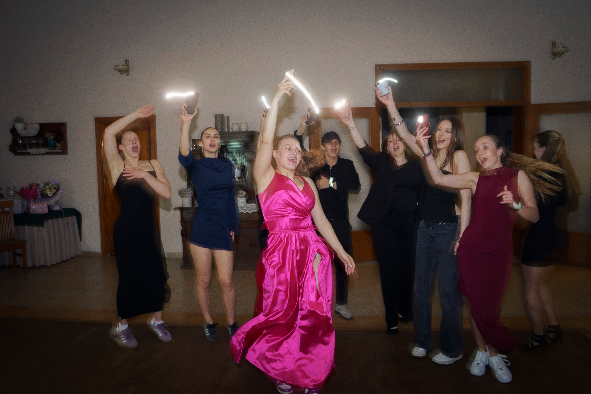 Rozbawiona grupa młodych ludzi tańczy z telefonami w górze, oświetlając salę. Uśmiechy, energia i swobodna atmosfera imprezy.