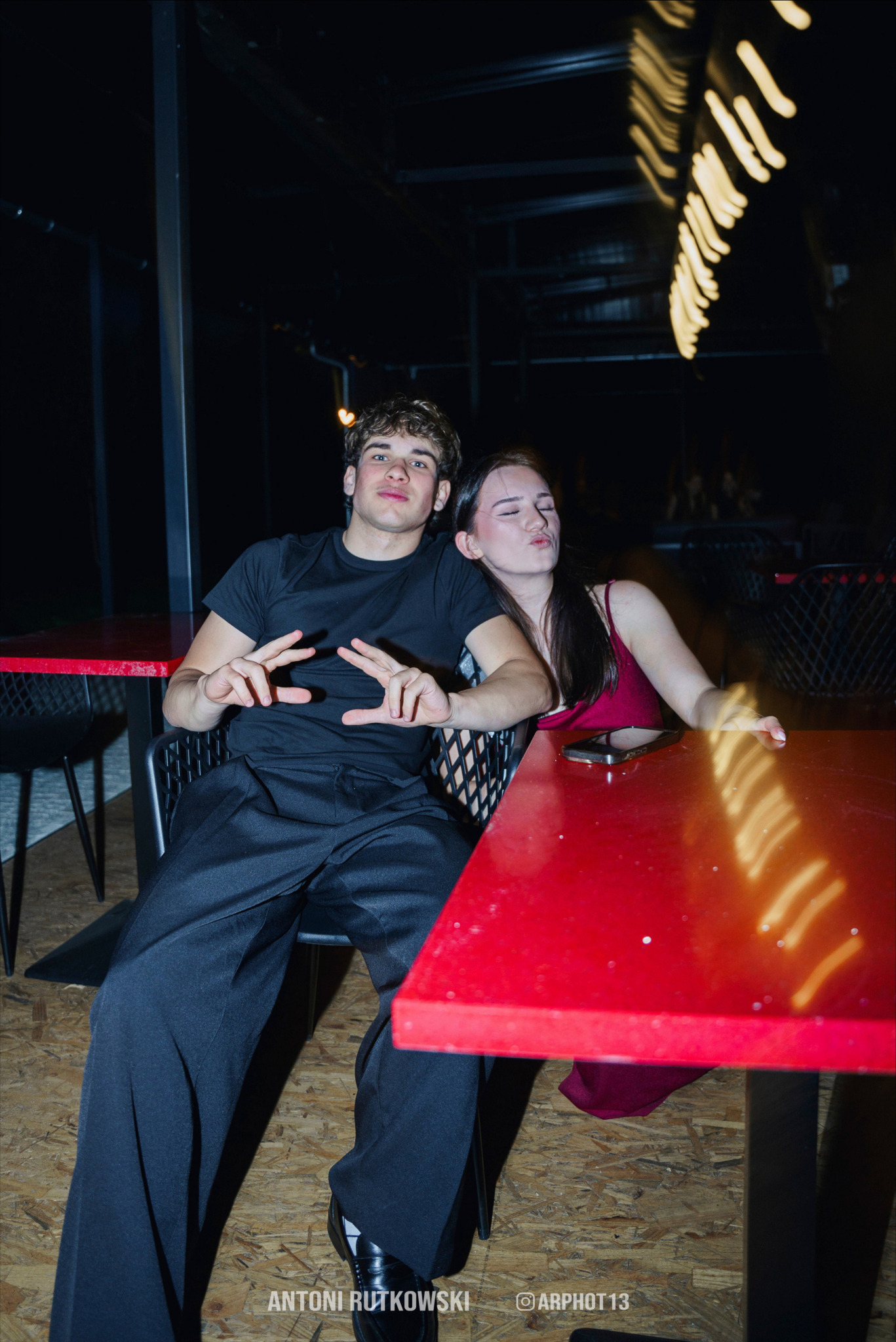 Młody mężczyzna i kobieta siedzą przy czerwonym stole, mężczyzna pokazuje gest V, kobieta robi dzióbek, w tle widoczne dekoracyjne oświetlenie.