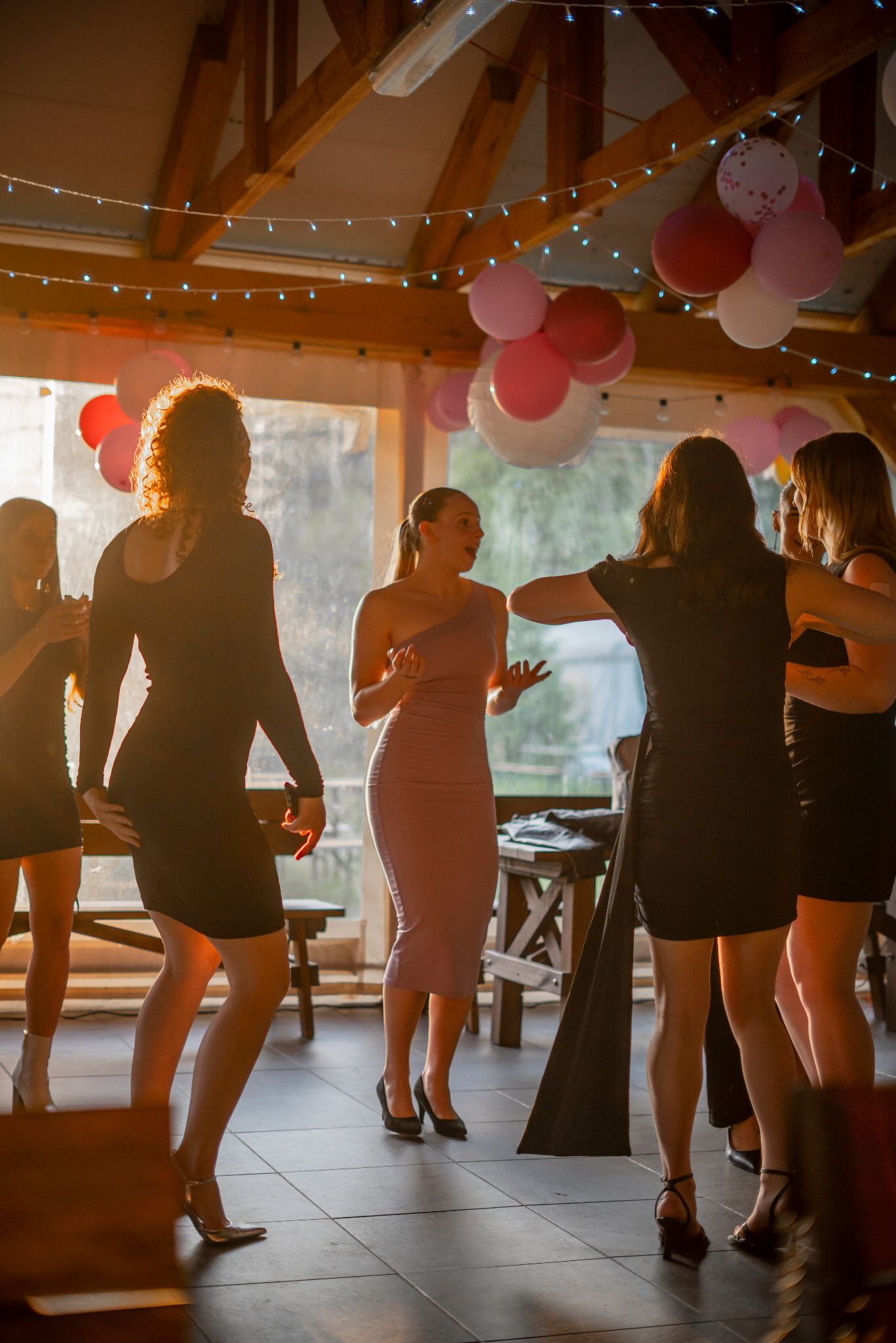 Radosna atmosfera na imprezie: grupa kobiet tańczy w eleganckich sukienkach pod drewnianą konstrukcją z balonami i lampkami, ciepłe światło podkreśla dynamikę zabawy.