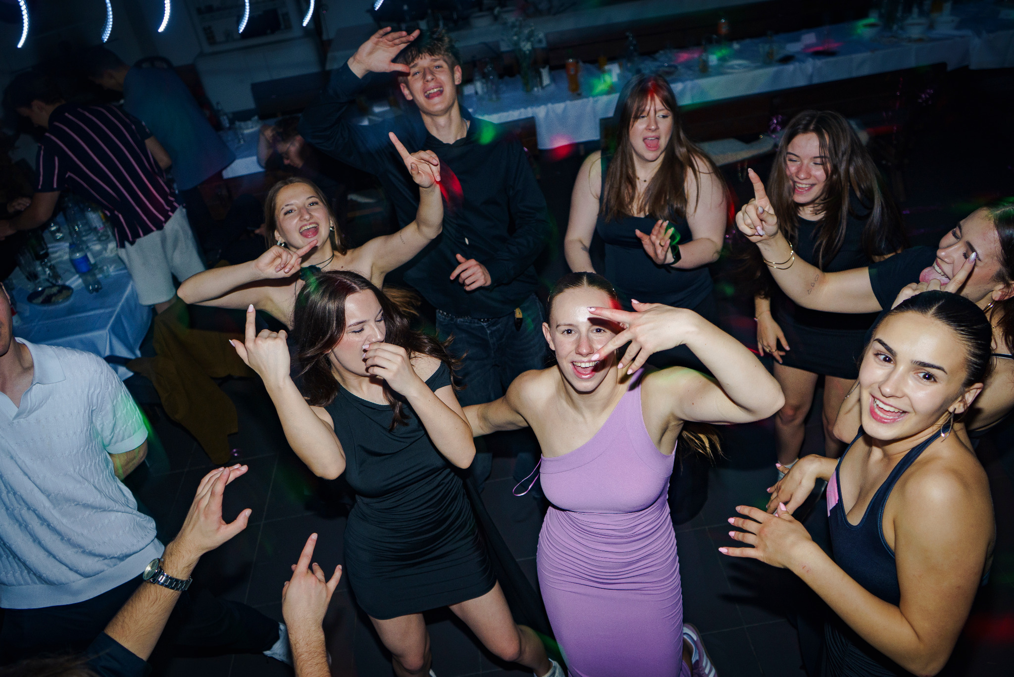 Radosna grupa młodych ludzi tańczy i pozuje podczas imprezy. Zdjęcie wykonane w Opolu, z dynamicznym oświetleniem i uśmiechniętymi twarzami.