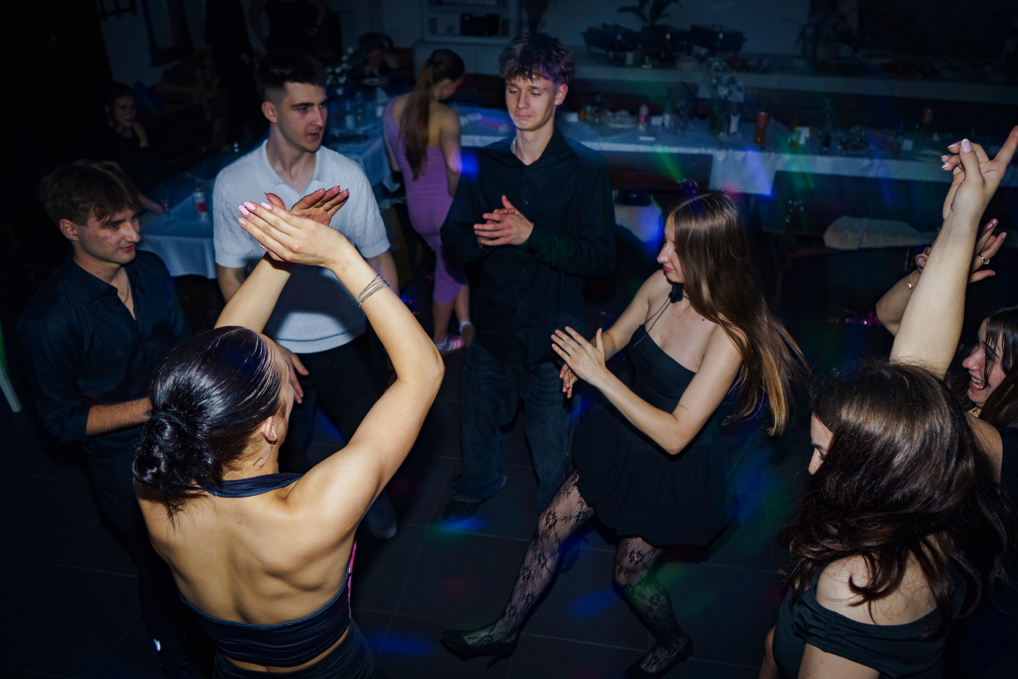 Energetyczne ujęcie z imprezy: grupa młodych ludzi tańczy w rytm muzyki, oświetleni kolorowymi światłami, tworząc dynamiczną i radosną atmosferę.