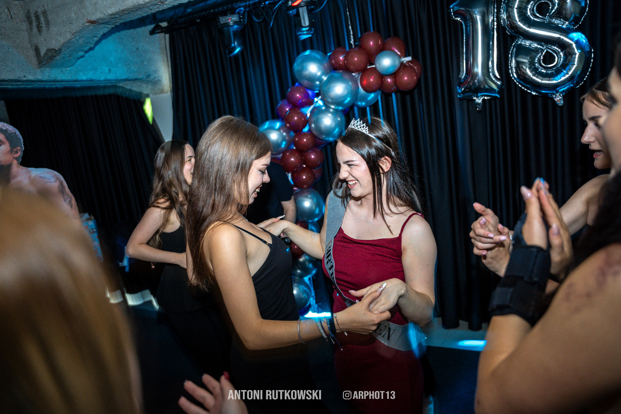 Uśmiechnięte nastolatki świętują 18 urodziny, z balonami i dekoracjami na tle czarnych zasłon. Jedna z dziewcząt ma koronę i szarfę 'Birthday Girl'.